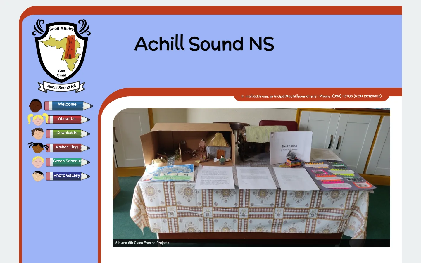 The existing Achill Sound NS homepage on a desktop screen: a light blue two-column layout with a crest, pencil-shaped menu buttons and one large photo of a classroom project